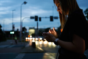 Close up shot of woman using smartphone on city street with bokeh lights at night. Mobile phone in female hands outdoors - obrazy, fototapety, plakaty