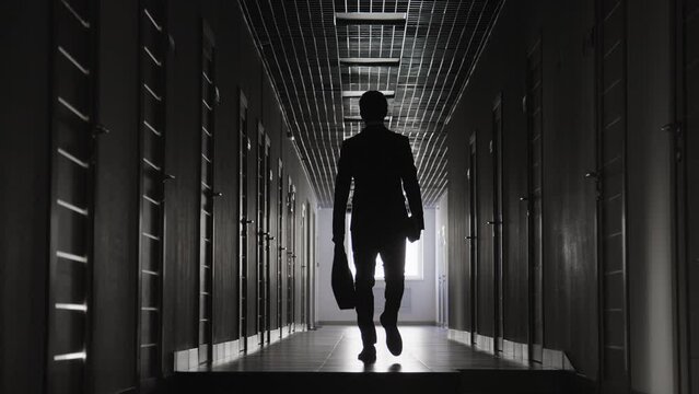 View from behind of unrecognizable male figure wearing formal suit in dark corridor carrying briefcase while walking away