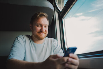 A man with a beard and mustache in a blue t-shirt is using a smartphone while traveling by Railway...