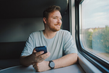 A man with a beard and mustache in a blue t-shirt is using a smartphone while traveling by Railway...