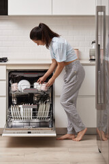 happy woman unloading plate from open automatic built-in dishwasher machine with clean utensils...