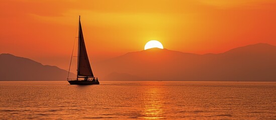 A sailboat with raised sails is outlined against the sunset and hazy orange sky amidst two coastal landmasses in Costa Rica copy space image - obrazy, fototapety, plakaty