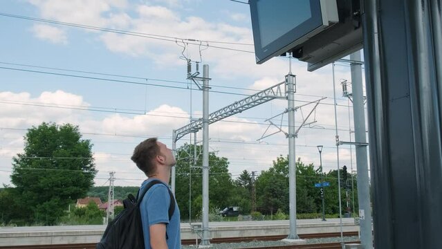 Man backpacker on the train station looking at train schedule display. Male tourist worried about the train delay and looks at the information on the board. Travel by railroad concept