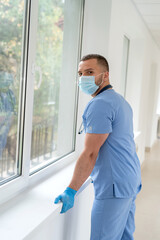 Male caucasian doctor wearing blue medical coat and face mask in modern clinic corridor