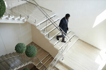 High angle view at Black man in business suit going downstairs holding on handrail in modern office...