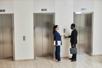 Wide angle view at Caucasian businesswoman with cup and laptop talking to cheerful African American businessman with phone and briefcase dressed in suits standing by elevators in lobby, copy space