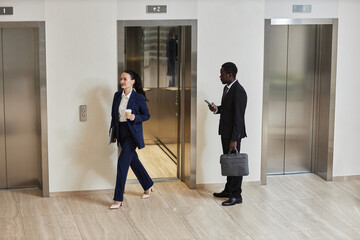 Wide shot of African American businessman holding phone and bag looking at Caucasian business lady exiting elevator in office building - Powered by Adobe