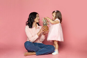 Young brunette woman and baby girl holding fruit on pink background