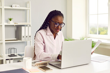 Female accountant at work. Young African American woman in glasses with Afro braids sitting at office desk with cellphone, coffee and calculator working on laptop computer and doing business paperwork