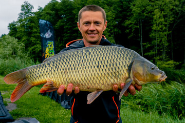 portrait of a satisfied professional fisherman holding a carp on the bank of a river fishing in a pond with a good catch