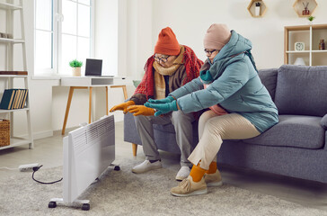 Portrait of a senior frozen elderly couple sitting on the sofa in the living room in winter outerwear and hats at home and trying to warm on electric heater. Heating problems concept.