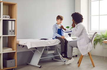 Friendly doctor doing medical checkup of child patient. Young African American woman pediatrician in white coat puts stethoscope to chest of happy little boy and listens to his heart or lungs sounds - obrazy, fototapety, plakaty