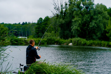 Fisherman with fishing rod or spinning and professional tools sitting on the river bank rear view...