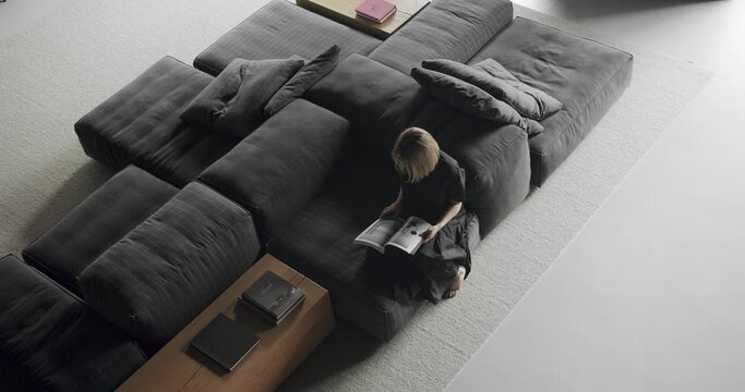 Overhead view of woman sitting on comfort modular sofa and reading magazine at modern apartment. Lounge zone for rest and relax in living room with loft style