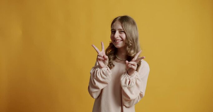 Funny blonde kid teen teenager girl 13 years in a beige dress she is happy and smiling and showing victory sign on a yellow background. teenage girl smiling and show two fingers, victory gesture.