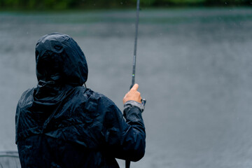 Fisherman in raincoat with fishing rod or spinning and professional tools sitting on river bank...