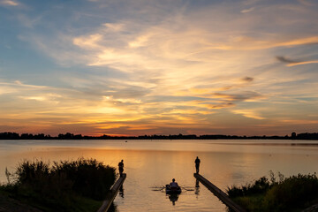 One angler in a boat and two on the shore during a beautiful sunset over lake Zoetermeerse Plas - obrazy, fototapety, plakaty