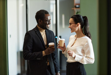 Waist up shot of two ethnically diverse coworkers laughing and drinking coffee having a discussion while standing inside modern office building
