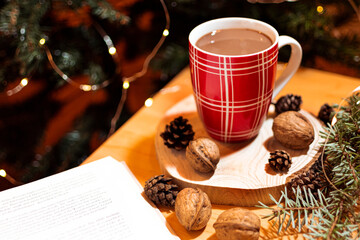 A cosy Christmas ambience featuring a cup of rich cocoa resting on a rustic wooden plate surrounded by pine cones and walnuts with a Christmas tree in the background and an open book next to the cup