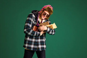 Smiling young man in checkered jacket, warm hat and sunglasses holding firewood against green studio background. Concept of human emotions, youth, winter fashion, vacations holidays