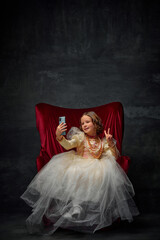 Little princess, girl sitting on royal throne and taking selfie showing piece sign against dark vintage background.