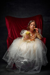 Little princess eating, tasting hot fries potato at fist time sitting in royal throne against dark...