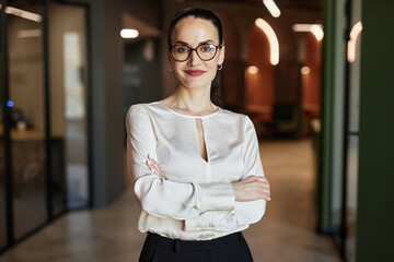 Waist up portrait of smiling Caucasian brunette businesswoman in glasses crossing arms and looking...