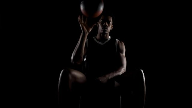 Basketball player balancing a ball. African american sports man sitting and practicing against black background.