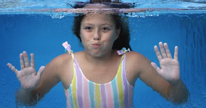 Super slow motion of little girl in colorful swimsuit having fun to swim underwater and greet with hand splashing water in transparent swimming pool during summer holidays travel vacation on sunny day