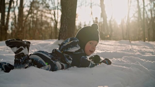 A cheerful child is happily lying around in a snowdrift in the forest. The boy is happy about the first snow and the arrival of winter, he plays and has fun in nature.