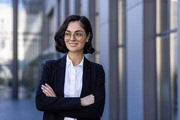 Fototapeta na wymiar Portrait of a young business woman in glasses and a suit standing outside an office building with her arms crossed on her chest. He looks away with a smile, a close-up photo