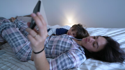 Mother and child laid in bed staring at cellphone device screen. Parent holding phone with one arm...