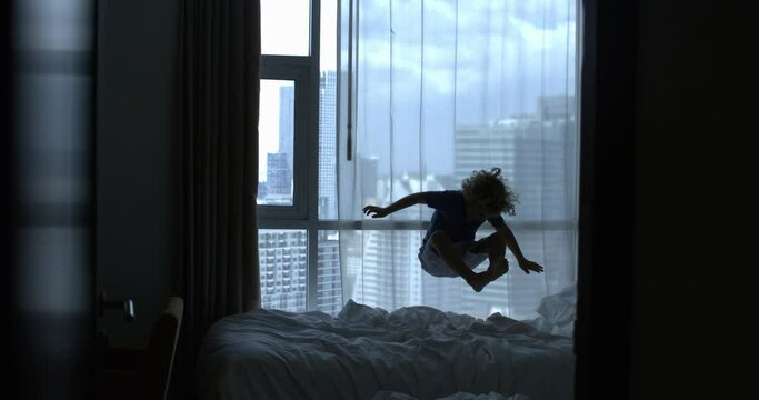 Super slow motion of carefree little boy is having fun to jump on bed at home in complete freedom and happiness in hotel room with view of skyscrapers during holidays family vacation trip.