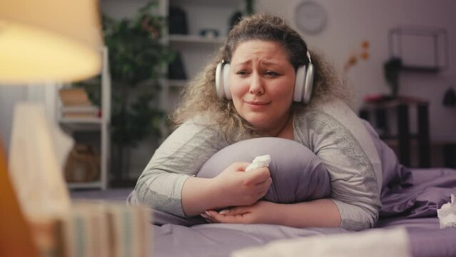 Sad plus-size woman crying on bed, listening to sentimental music in earphones