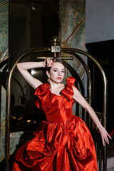 Fototapeta na wymiar Pretty woman in a luxurious red dress posing in a luggage trolley.Fashion shooting concept