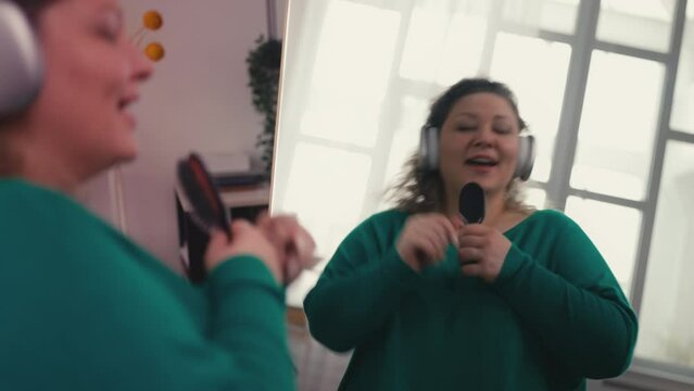 Pretty overweight woman listening music in earphones, singing with hair brush