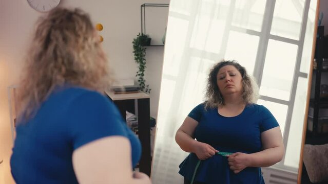 Sad overweight woman measuring waist in front of mirror, unhappy with result