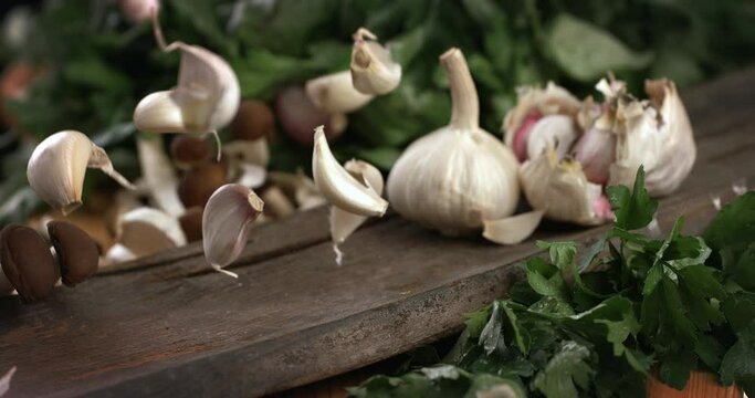 Slow motion macro of falling fresh ripe organic garlic cloves on composition with bulbs and parsley on wooden cut board in kitchen of restaurant.