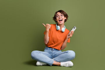 Full size photo of excited woman wear earphones sit look directing empty space smartphone in hand...