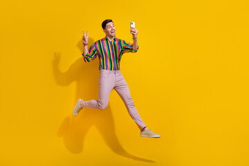 Fototapeta na wymiar Full size photo of young guy trampoline jumper show v sign at party hipsters striped shirt make selfie isolated on yellow color background