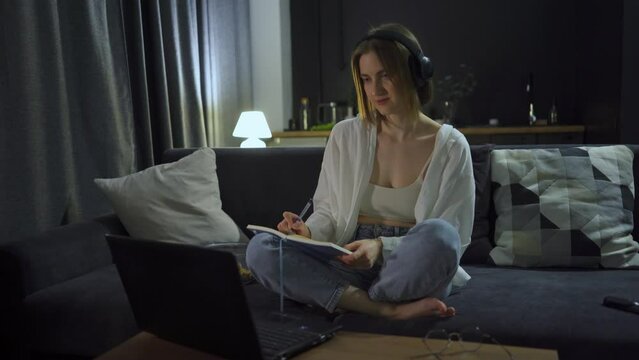 Young student girl wearing headphones in the evening takes notes on an online lecture, conducts a video conference, and studies a foreign language. Caucasian woman is learning a new profession online - Powered by Adobe