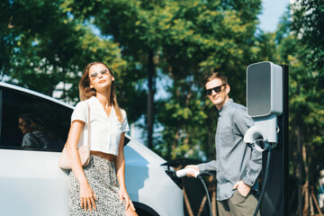 Young couple travel with EV electric car charging in green sustainable city outdoor garden in...