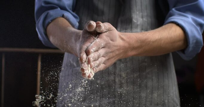 Super slow motion macro of professional artisan baker chef claps his hands with flour while prepares a dough for making homemade bread, pasta or pizza on rustic wooden table in traditional bakery. 