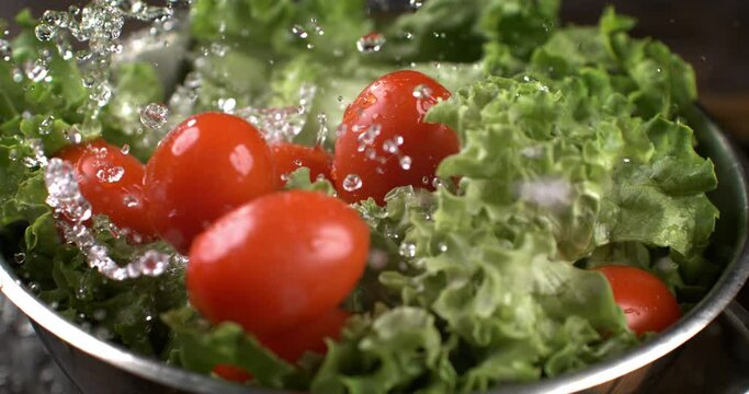 Super slow motion macro of splashing water drops are falling on fresh ripe organic tomatoes and green salad while being washed in colander in kitchen of restaurant.