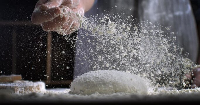 Super slow motion close up of professional artisan baker chef sprinkles flour on raw loaf of dough while making homemade bread, pasta or pizza on rustic wooden table in traditional bakery kitchen.