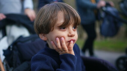 Pensive child thinking while seated on stroller with hand in chin. Young boy observing world with thoughtful expression while being carried in carriage