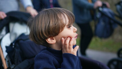 Pensive child thinking while seated on stroller with hand in chin. Young boy observing world with thoughtful expression while being carried in carriage