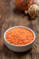 A high angle close-up image of a bowl of red lentils on a vintage wooden table with onion and garlic in the background