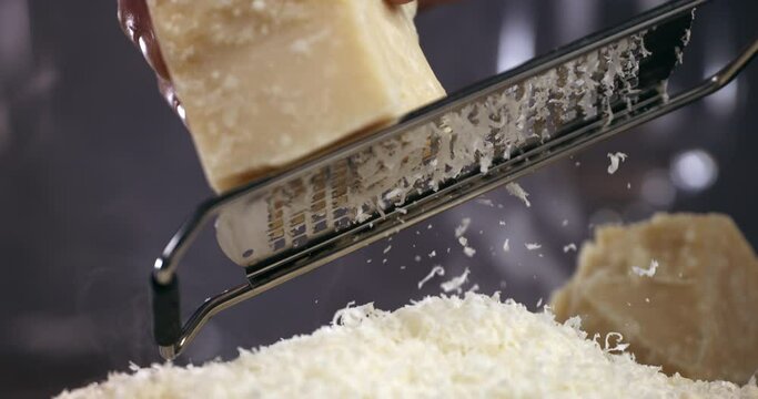 Super slow motion macro of a professional chef grating traditional parmesan cheese with steel grater on the dish close up and pieces of cheese fall at 1000 fps for preparing pasta.