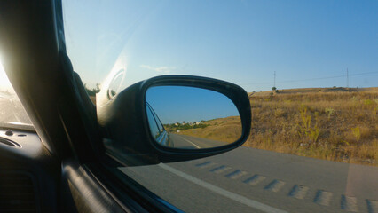 Fototapeta na wymiar View from the car in the side mirror. A car is driving on a tarmac road in the countryside. There are yellow fields on the side of the road. The mirror reflects the road and the blue sky.